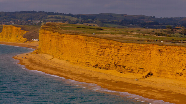Aerial view of the golden sandstone cliffs of the Jurassic Coast meeting the sea at West Bay with gentle waves washing onto the sandy beach West Bay England United Kingdom. Aerial view of the golden sandstone cliffs of the Jurassic Coast meeting the sea at West Bay with gentle waves washing onto the sandy beach West Bay England United Kingdom.