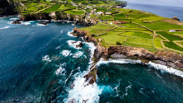Aerial view of verdant fields meeting the turquoise ocean near Estrada do Farol where foamy waves crash against the rugged cliffs Flores A&ccedil;ores Portugal.
