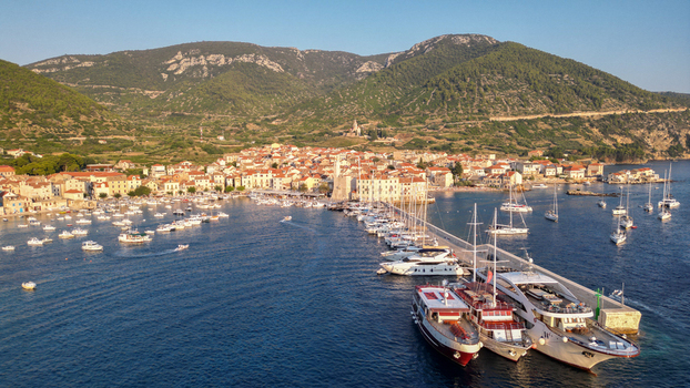 Aerial view of Komiza harbor with boats moored at the stone pier historic buildings and lush green mountains under a clear sky Komiza Split Dalmatia County Croatia.