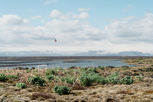 View of a person kitesurfing on a river with mossy volcanic terrain in the foreground and snow capped mountains in the distance in Skaftarhreppur Iceland.
