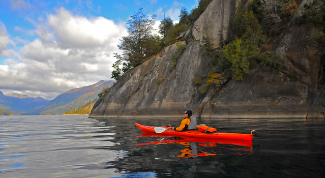 View of a man in a red kayak paddling on the calm waters of Nahuel Huapi Lake by steep rocky cliffs San Carlos de Bariloche Rio Negro Argentina. View of a man in a red kayak paddling on the calm waters of Nahuel Huapi Lake by steep rocky cliffs San Carlos de Bariloche Rio Negro Argentina.