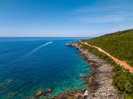 Aerial view of Aquarium Beach featuring turquoise waters rocky coastline a boat leaving a wake and a dirt road winding through lush green hills in Jala Vlore County Albania.