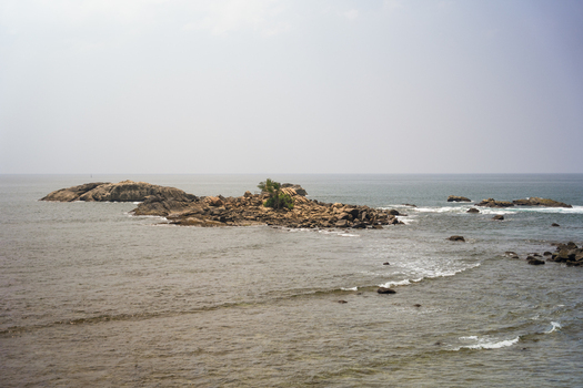 View of a rocky islet with a small green tree surrounded by gentle ocean waves under a hazy sky in Galle Southern Province Sri Lanka.