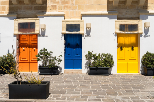 View of three vibrant orange blue and yellow wooden doors on a white facade with potted green plants and stone paving in Marsaskala Southern Region Malta. View of three vibrant orange blue and yellow wooden doors on a white facade with potted green plants and stone paving in Marsaskala Southern Region Malta.