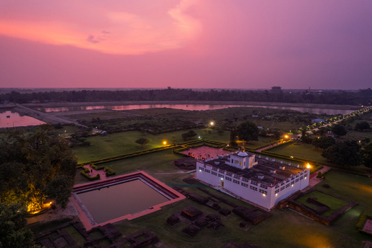 Aerial view of the Maya Devi Temple in Lumbini featuring the sacred pond ancient ruins and illuminated temple structure under a vibrant sunset sky Nepal.
