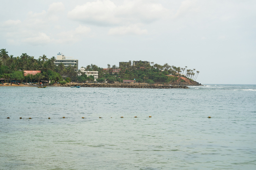 View of the tropical coastline with palm trees beachfront buildings and a stone breakwater under a cloudy sky in Mirissa Southern Province Sri Lanka. View of the tropical coastline with palm trees beachfront buildings and a stone breakwater under a cloudy sky in Mirissa Southern Province Sri Lanka.