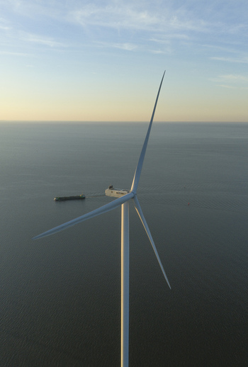 Aerial view of a large offshore wind turbine standing tall over the calm sea with a cargo ship sailing in the distance during golden hour in Netherlands. Aerial view of a large offshore wind turbine standing tall over the calm sea with a cargo ship sailing in the distance during golden hour in Netherlands.