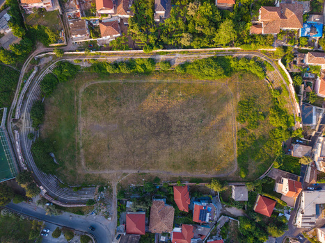 Aerial view of a small football stadium with an overgrown pitch and running track surrounded by residential houses and green trees in Kruje Durres County Albania.
