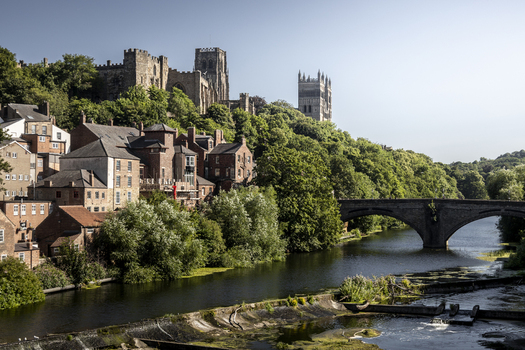 View of Durham Cathedral and Durham Castle overlooking the River Wear with lush green trees and a stone bridge in the foreground in Durham England United Kingdom. View of Durham Cathedral and Durham Castle overlooking the River Wear with lush green trees and a stone bridge in the foreground in Durham England United Kingdom.