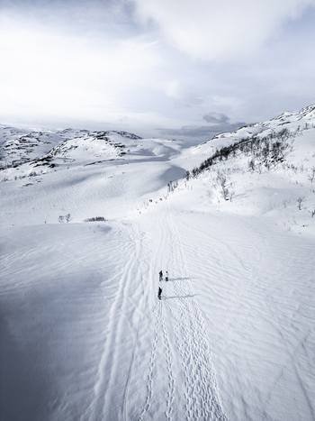 Aerial view of skiers walking on a snowy path through a vast mountain landscape under a cloudy sky Kvaloya Troms Norway. Aerial view of skiers walking on a snowy path through a vast mountain landscape under a cloudy sky Kvaloya Troms Norway.