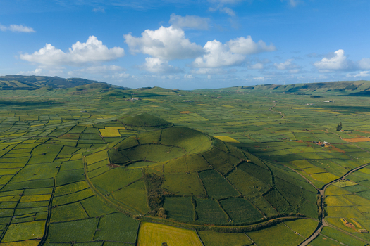 Aerial view of Pico Dona Joana volcanic crater surrounded by a patchwork of green agricultural fields and stone walls under a blue sky in Feteira Acores Portugal.