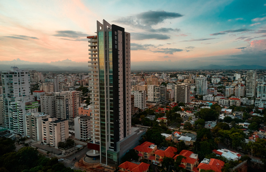Aerial view of a modern skyscraper rising above the urban skyline at sunset surrounded by residential buildings near Mirador Sur Santo Domingo Distrito Nacional Dominican Republic.