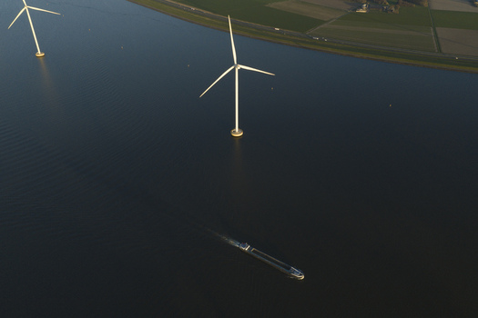 Aerial view of offshore wind turbines standing in calm water with a cargo ship cruising past green fields at sunset in Netherlands. Aerial view of offshore wind turbines standing in calm water with a cargo ship cruising past green fields at sunset in Netherlands.