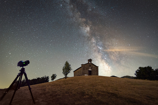 View of the stone church at Passo di Cirone under the Milky Way galaxy and a starry night sky with a camera on a tripod in the foreground in Pontremoli Toscana Italy. View of the stone church at Passo di Cirone under the Milky Way galaxy and a starry night sky with a camera on a tripod in the foreground in Pontremoli Toscana Italy.
