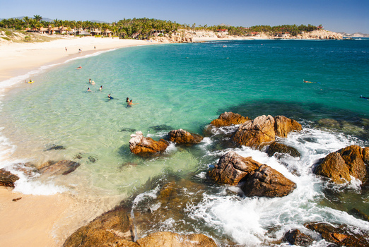 View of Chileno Bay Public Beach with turquoise water rocky foreground and people swimming under a clear blue sky in Cabo San Lucas Baja California Sur Mexico. View of Chileno Bay Public Beach with turquoise water rocky foreground and people swimming under a clear blue sky in Cabo San Lucas Baja California Sur Mexico.