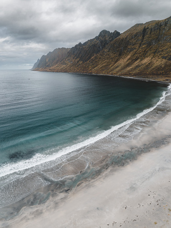 Aerial view of the dramatic coastline with rugged mountains meeting the dark ocean and a sandy beach under a cloudy sky in Senja Troms Norway.