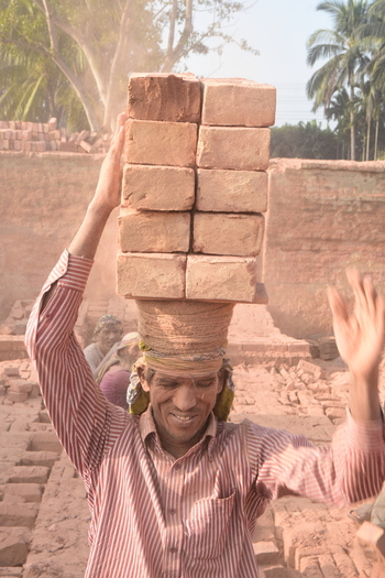 Kushtia District Khulna Division Bangladesh 28 November 2020 View of a smiling worker carrying a heavy stack of red bricks on his head at a dusty brickyard. Kushtia District Khulna Division Bangladesh 28 November 2020 View of a smiling worker carrying a heavy stack of red bricks on his head at a dusty brickyard.