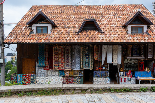 View of traditional wooden house with terracotta tiled roof and colorful handmade rugs displayed at a local shop with a vendor sitting inside in Kruje Durres County Albania.