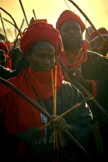 Zaria Nigerian 21 March 2026 View of intense warriors in vibrant red and deep shadows holding bows and arrows under a luminous golden sky. Zaria Nigerian 21 March 2026 View of intense warriors in vibrant red and deep shadows holding bows and arrows under a luminous golden sky.