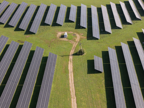 Aerial view of rows of solar panels in a large solar farm with a dirt path and electrical equipment on green grass in Poland.