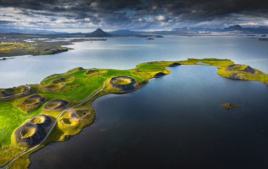 Aerial view of the Skutustadagigar pseudocraters on the shores of Lake Myvatn with green grassy hills and calm water under a dramatic cloudy sky Iceland.