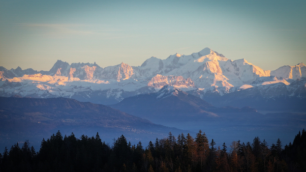 Aerial view of the snow capped peaks of the Mont Blanc massif glowing in the golden hour light above a dark pine forest and hazy blue valleys Chamonix Auvergne Rhone Alpes France. Aerial view of the snow capped peaks of the Mont Blanc massif glowing in the golden hour light above a dark pine forest and hazy blue valleys Chamonix Auvergne Rhone Alpes France.