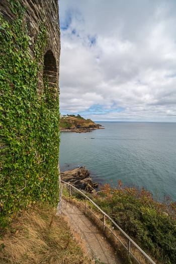 View of the Fowey River coastline with a stone wall covered in ivy a walking path rocky cliffs and distant buildings in Fowey Cornwall United Kingdom. View of the Fowey River coastline with a stone wall covered in ivy a walking path rocky cliffs and distant buildings in Fowey Cornwall United Kingdom.