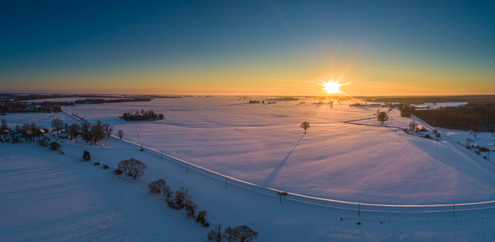 Aerial view of a vast snow covered field at sunrise with a winding road scattered trees and long shadows stretching across the frozen landscape in Latvia. Aerial view of a vast snow covered field at sunrise with a winding road scattered trees and long shadows stretching across the frozen landscape in Latvia.