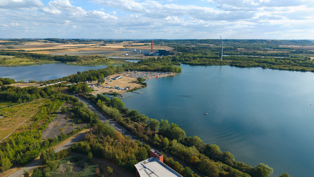 Aerial view of Stewartby Lake featuring calm blue water surrounding greenery a parking area with vehicles and a distant industrial facility in Bedford England United Kingdom. Aerial view of Stewartby Lake featuring calm blue water surrounding greenery a parking area with vehicles and a distant industrial facility in Bedford England United Kingdom.