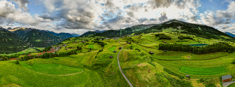 Aerial view of the village of Prutz in the Tyrolean Alps featuring lush green meadows wooden barns a winding road and mountain peaks under a cloudy sky in Prutz Tyrol Austria. Aerial view of the village of Prutz in the Tyrolean Alps featuring lush green meadows wooden barns a winding road and mountain peaks under a cloudy sky in Prutz Tyrol Austria.