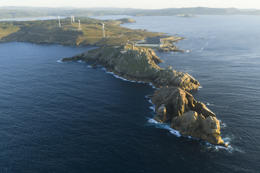 Aerial view of a rocky peninsula featuring a lighthouse surrounded by the Atlantic Ocean with wind turbines on the distant coastline in Galicia Spain.