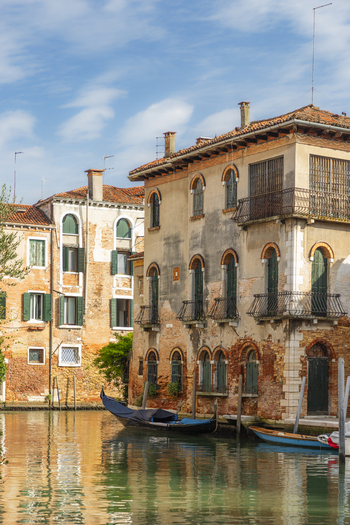 View of a traditional gondola moored on a quiet canal in front of historic brick buildings with green shutters in Venice Veneto Italy. View of a traditional gondola moored on a quiet canal in front of historic brick buildings with green shutters in Venice Veneto Italy.