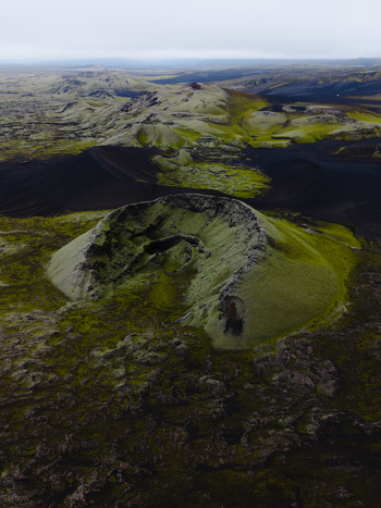 Aerial view of a moss covered volcanic cone crater rising from a dark rugged landscape under a pale sky Iceland. Aerial view of a moss covered volcanic cone crater rising from a dark rugged landscape under a pale sky Iceland.
