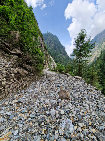 View of a rugged rocky mountain path surrounded by steep cliffs lush green forests and towering mountains under a bright blue sky Malana Himachal Pradesh India.