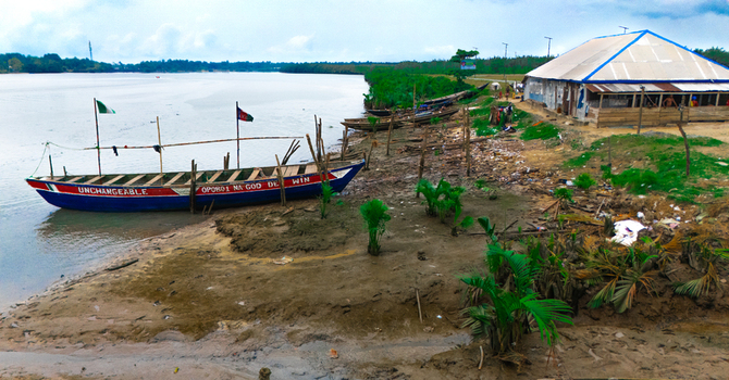 Aerial view of a wooden boat moored on the muddy bank of a river near a simple building with a corrugated metal roof in Minima Rivers Nigeria.