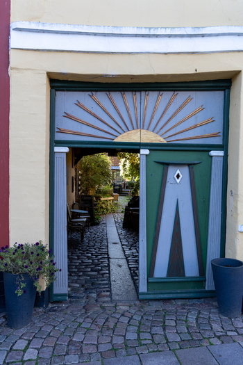 View of a decorative green wooden gate with a sunburst motif leading to a cobblestone courtyard with outdoor seating and lush greenery in Ribe Denmark. View of a decorative green wooden gate with a sunburst motif leading to a cobblestone courtyard with outdoor seating and lush greenery in Ribe Denmark.