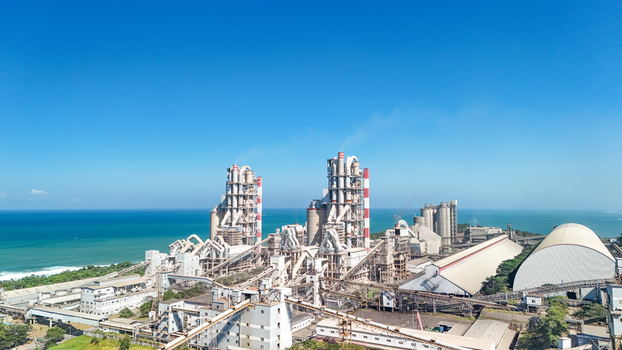 Aerial view of a large industrial cement plant with tall silos conveyor systems and a white dome structure situated along the coastline in Indonesia.