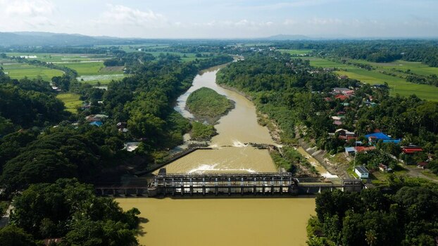 Aerial view of Jalaur Dam spanning the river surrounded by lush green vegetation agricultural rice fields and rural houses in Dingle Western Visayas Philippines.