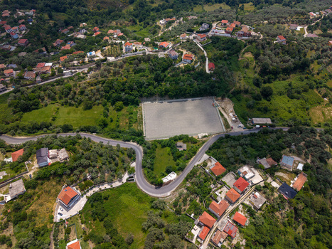 Aerial view of a hillside village with red roofed houses a winding road and a large rectangular construction site surrounded by lush green trees in Kruje Durres County Albania.