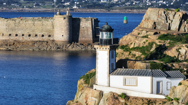 Aerial view of the stark white lighthouse juxtaposed against rugged cliffs and the imposing Ch&acirc;teau du Taureau fortress embraced by the deep blue sea Carantec Brittany France.