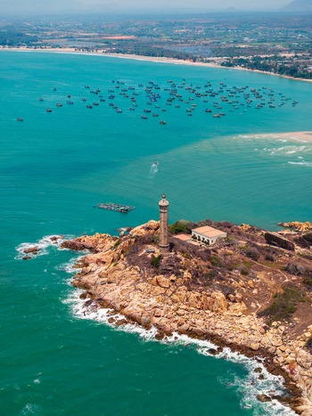 Aerial view of Ke Ga Lighthouse standing on a rocky peninsula surrounded by turquoise sea and numerous small fishing boats in the bay of Vietnam. Aerial view of Ke Ga Lighthouse standing on a rocky peninsula surrounded by turquoise sea and numerous small fishing boats in the bay of Vietnam.