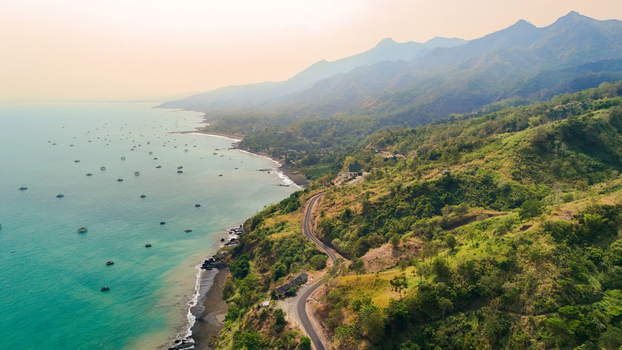 Aerial view of a winding coastal road along lush green hillsides overlooking the turquoise ocean filled with small fishing boats in Sukabumi Indonesia. Aerial view of a winding coastal road along lush green hillsides overlooking the turquoise ocean filled with small fishing boats in Sukabumi Indonesia.