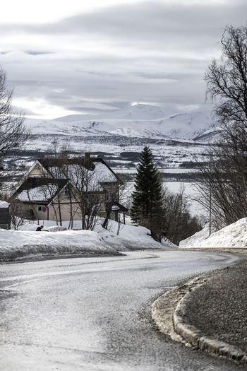 View of a winding icy road leading past snow covered houses towards distant white mountains under a cloudy sky in Tromso Troms Norway.
