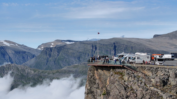 Aerial view of the Utsikten viewpoint on a cliff edge with camper vans parked tourists walking on the platform and dramatic mountain peaks rising above a sea of clouds in Nordland Norway. Aerial view of the Utsikten viewpoint on a cliff edge with camper vans parked tourists walking on the platform and dramatic mountain peaks rising above a sea of clouds in Nordland Norway.