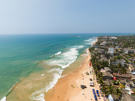 Aerial view of turquoise waves crashing on the sandy shore lined with palm trees and coastal buildings in Hikkaduwa Southern Province Sri Lanka. Aerial view of turquoise waves crashing on the sandy shore lined with palm trees and coastal buildings in Hikkaduwa Southern Province Sri Lanka.