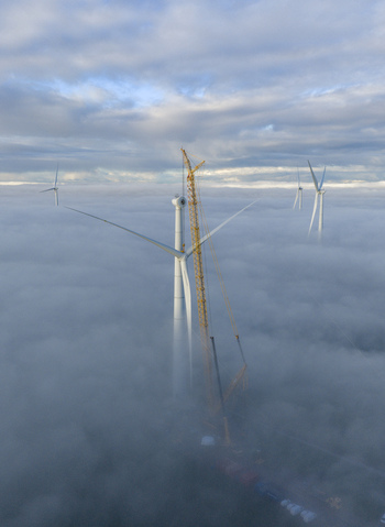 Aerial view of wind turbines and a yellow lattice boom crane emerging from a thick blanket of white fog under a cloudy sky.