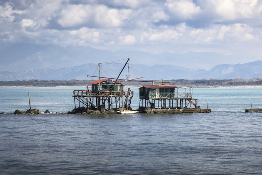View of traditional wooden fishing huts on stilts over the calm sea with mountains in the background Marina di Pisa Tuscany Italy.