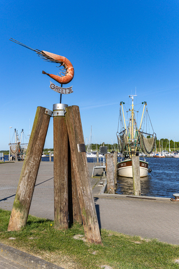 View of the Greetje shrimp sculpture on wooden pilings with a traditional fishing cutter in the harbor under a clear blue sky in Krummhorn Lower Saxony Germany. View of the Greetje shrimp sculpture on wooden pilings with a traditional fishing cutter in the harbor under a clear blue sky in Krummhorn Lower Saxony Germany.