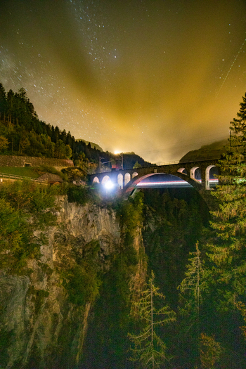 View of a stone railway viaduct illuminated at night with light trails from a passing train against a starry sky in the Swiss Alps Airolo Ticino Switzerland. View of a stone railway viaduct illuminated at night with light trails from a passing train against a starry sky in the Swiss Alps Airolo Ticino Switzerland.