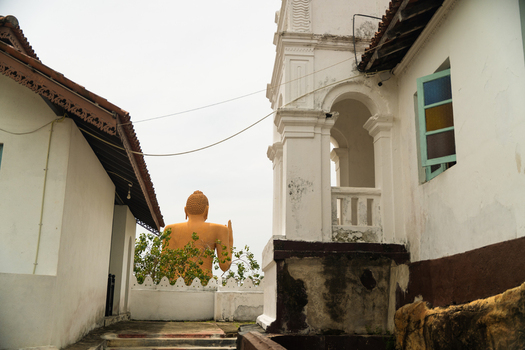 View of a large golden Buddha statue framed by traditional white temple buildings under an overcast sky in Dodanduwa Southern Province Sri Lanka. View of a large golden Buddha statue framed by traditional white temple buildings under an overcast sky in Dodanduwa Southern Province Sri Lanka.